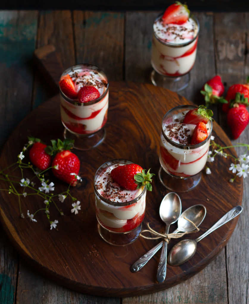 4 dessert glasses of strawberry tiramisu on a wooden board with silver spoons and white flowers.
