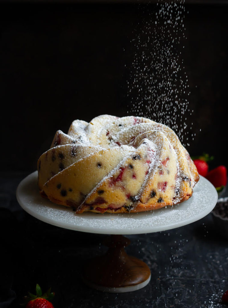 Powdered sugar getting sprinkled on cream cheese pound cake on a cake stand.
