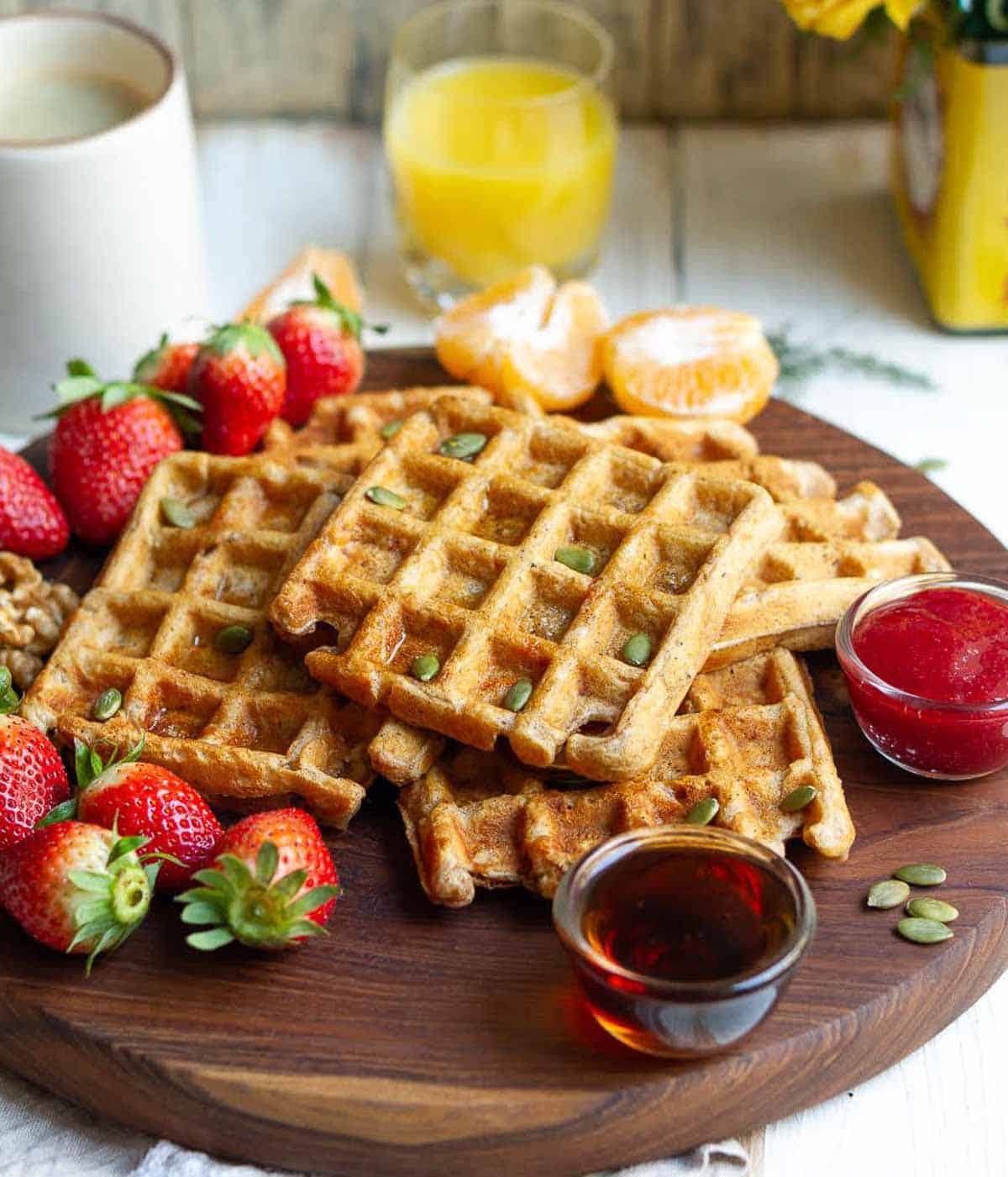 Waffles stacked on a wooden board with fruits around.