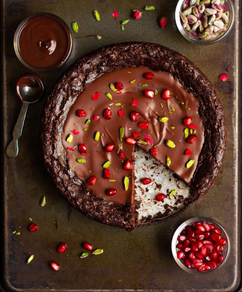 chocolate cake with ganache, pomegranates, and pistachios with a slice taken out.
