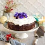 chocolate zucchini cake with cream cheese frosting and edible flowers on a cake stand.