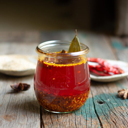 Chili oil in a glass jar on a wooden board with ingredients in the background.