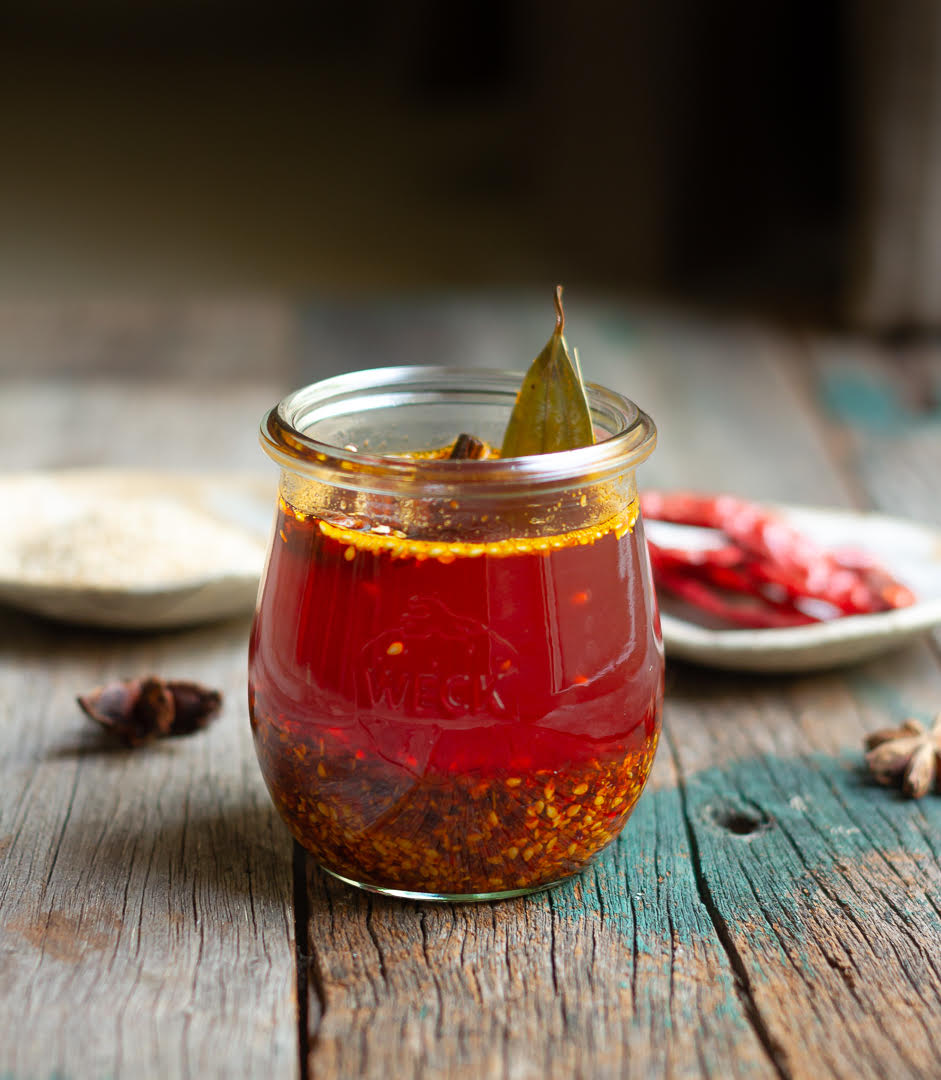 Chili oil in a glass jar on a wooden board with ingredients in the background.