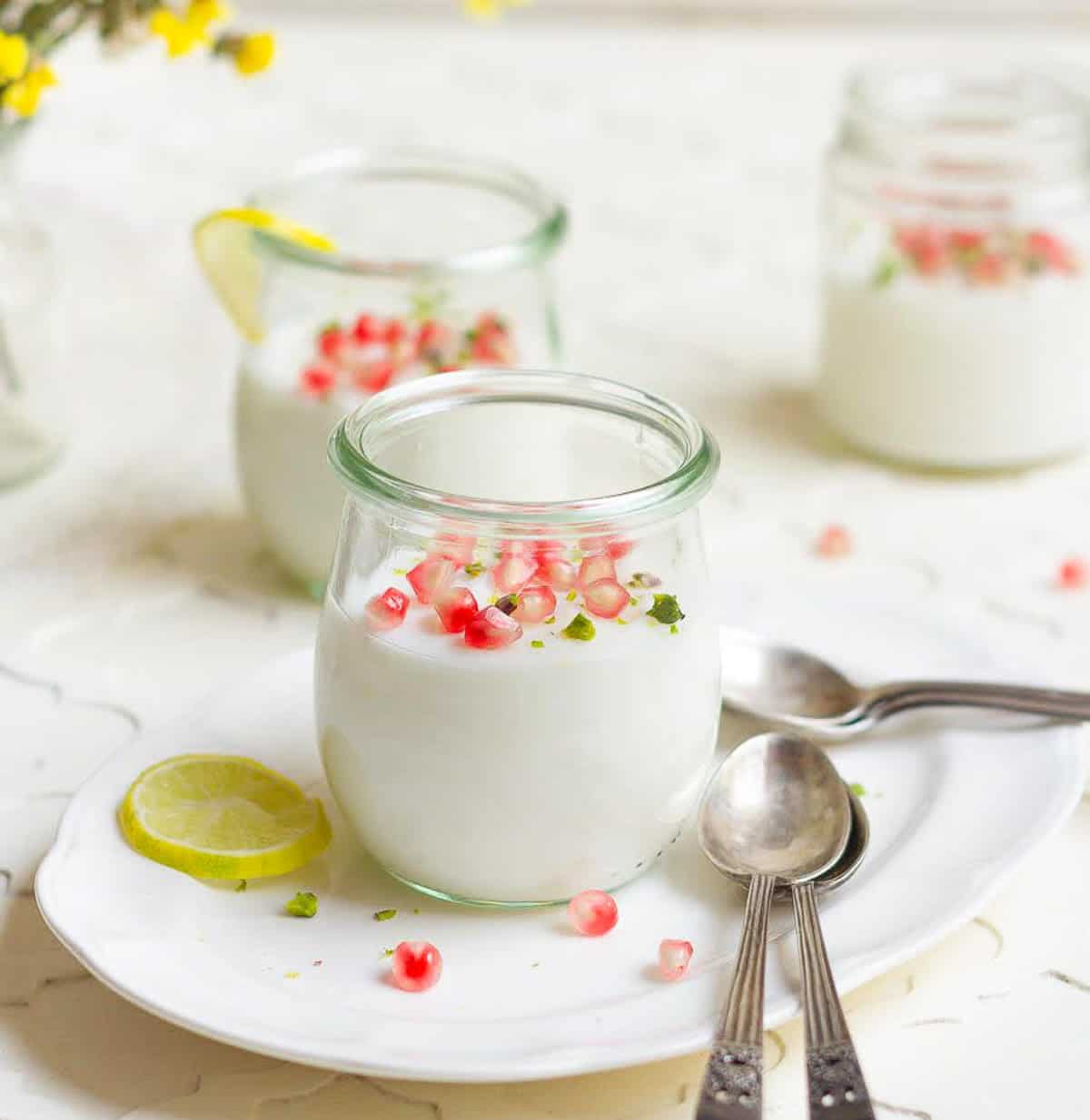 3 jars of panna cotta with pomegranates and lemon slices, yellow flowers, and spoons on the side.