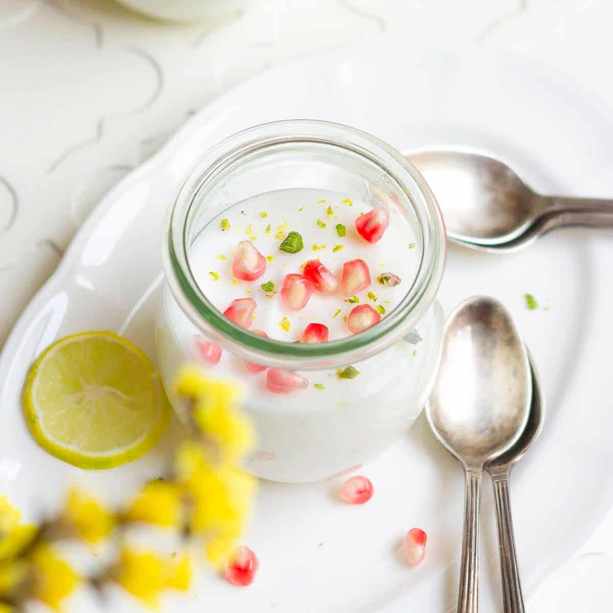 A jar of panna cotta garnished with pomegranate arils kept on a white platter with spoons.