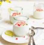 3 jars of panna cotta with yellow flowers, spoons, and lemon slice on the side.