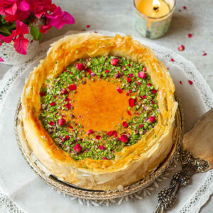 Baklava cheesecake garnished with pistachios and rose petals with flowers and a candle around.