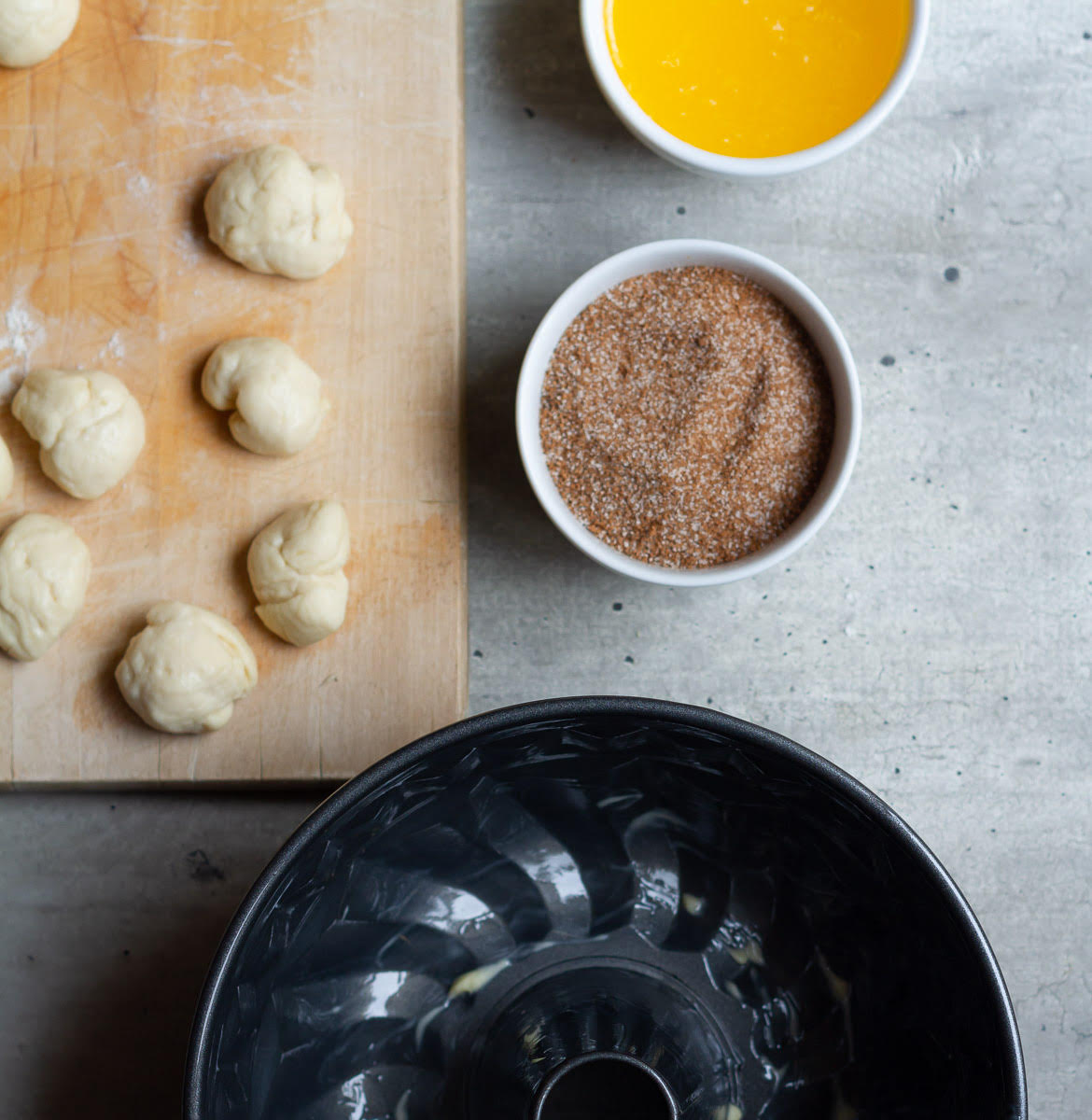 Dough pieces and a bundt pan with bowls of cinnamon sugar and melted butter.