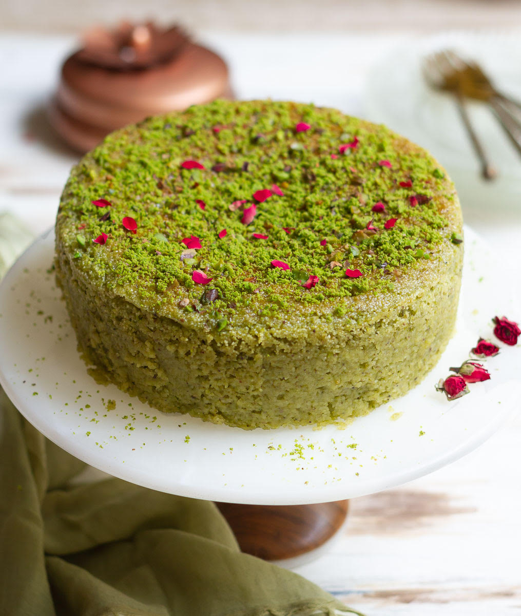Pistachio cake decorated with rose petals and pistachio powder on a cake stand.