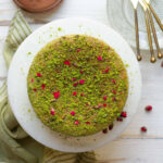 Pistachio cake topped with chopped pistachios and rose petals on a cake stand.