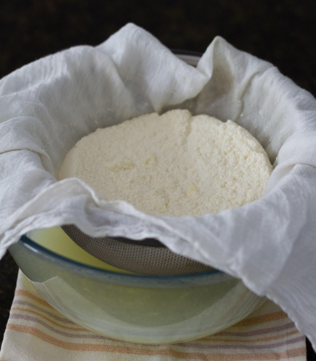 drained cheese curds in a cheesecloth lined sieve on a glass bowl.