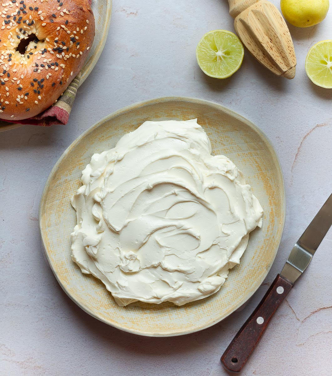 Cream cheese in a serving dish with a knife, lemon halves and bagels around it.
