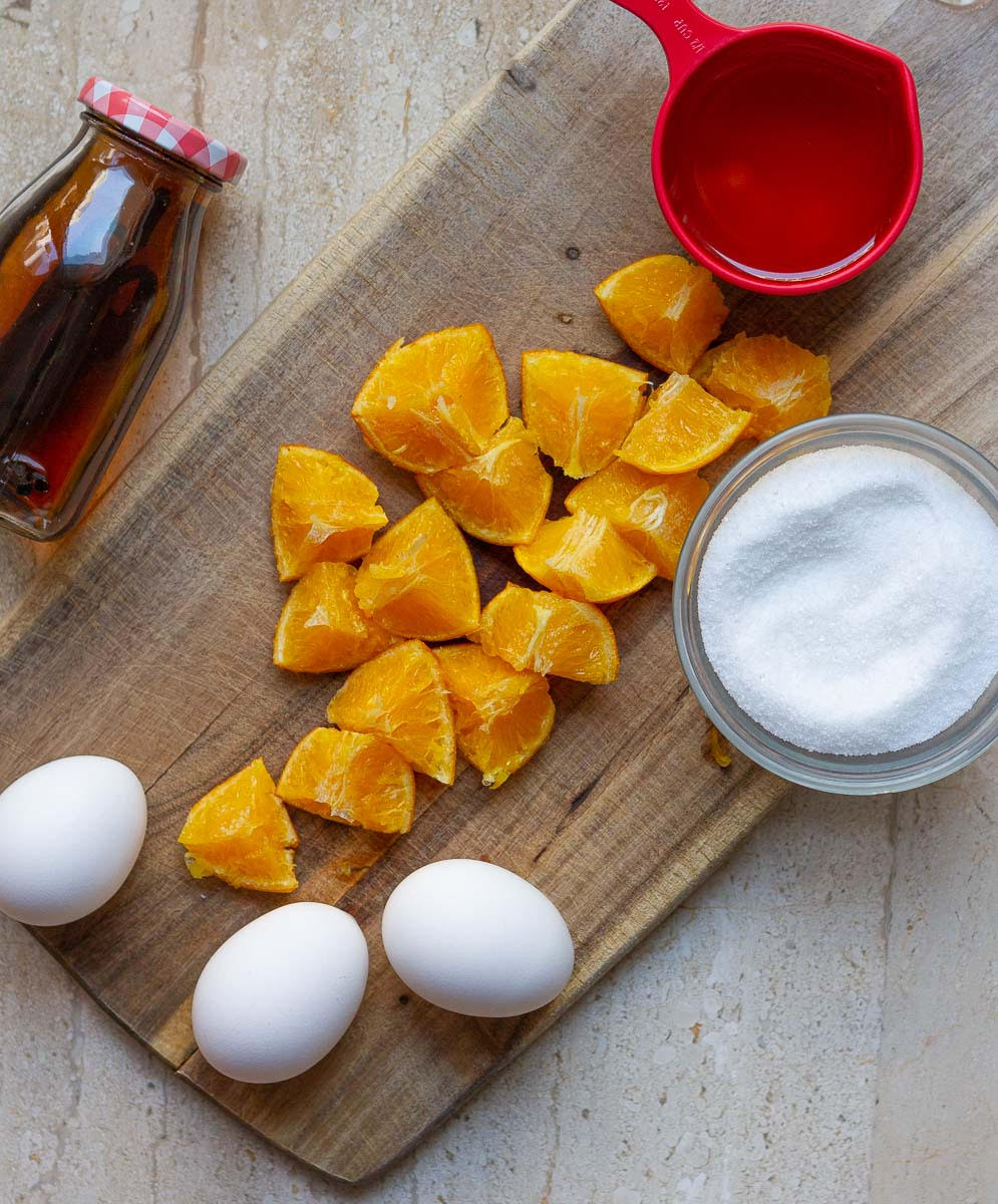 Cooked oranges, eggs, olive oil, sugar, vanilla extract on a wooden board.