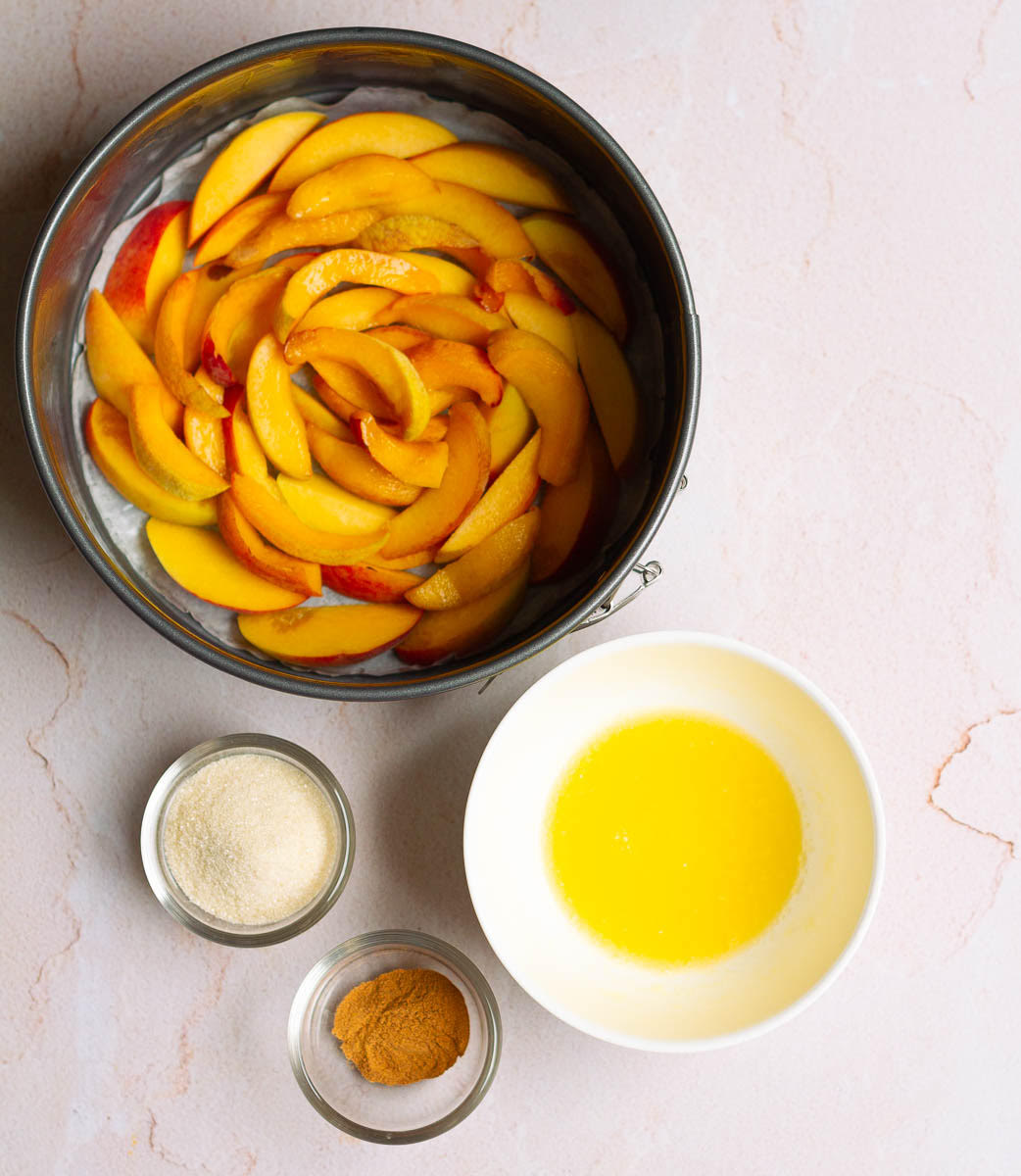 Sliced peaches lined at the bottom of cake pan and sugar, butter, cinnamon on the side.