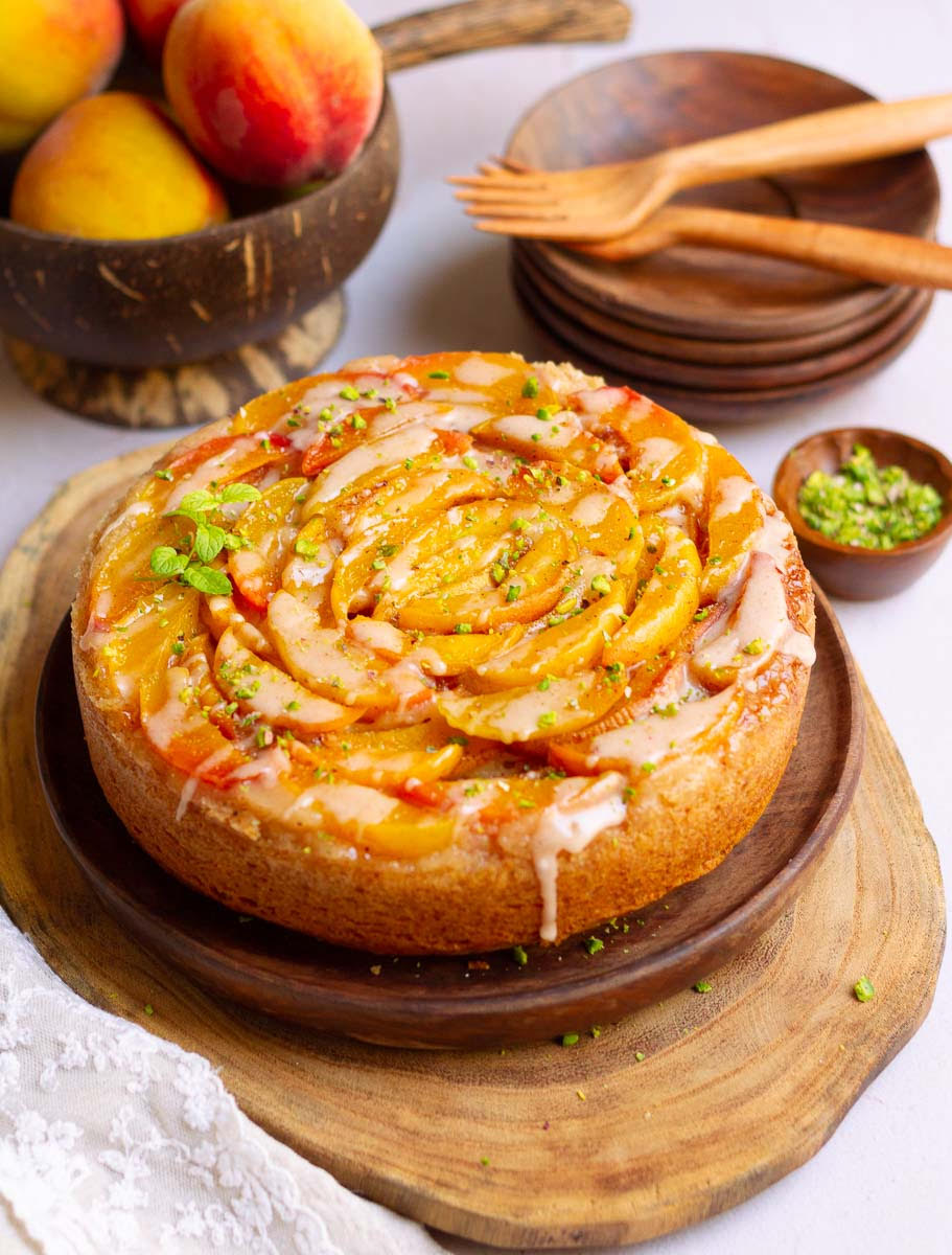 Peach cobbler cake on a wooden platter with peaches and plates in the background.