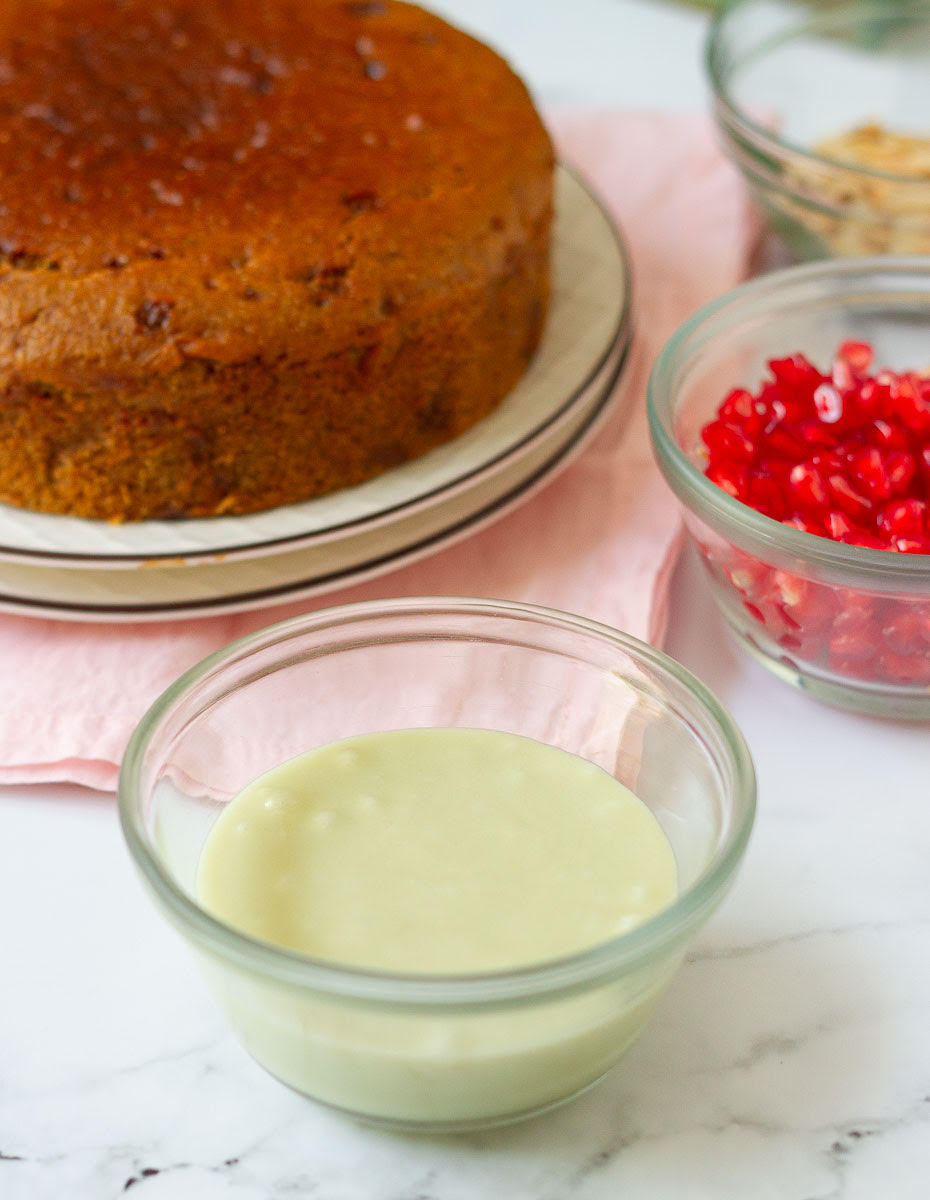 Cardamom glaze in a small bowl with cake and pomegranate in the background.