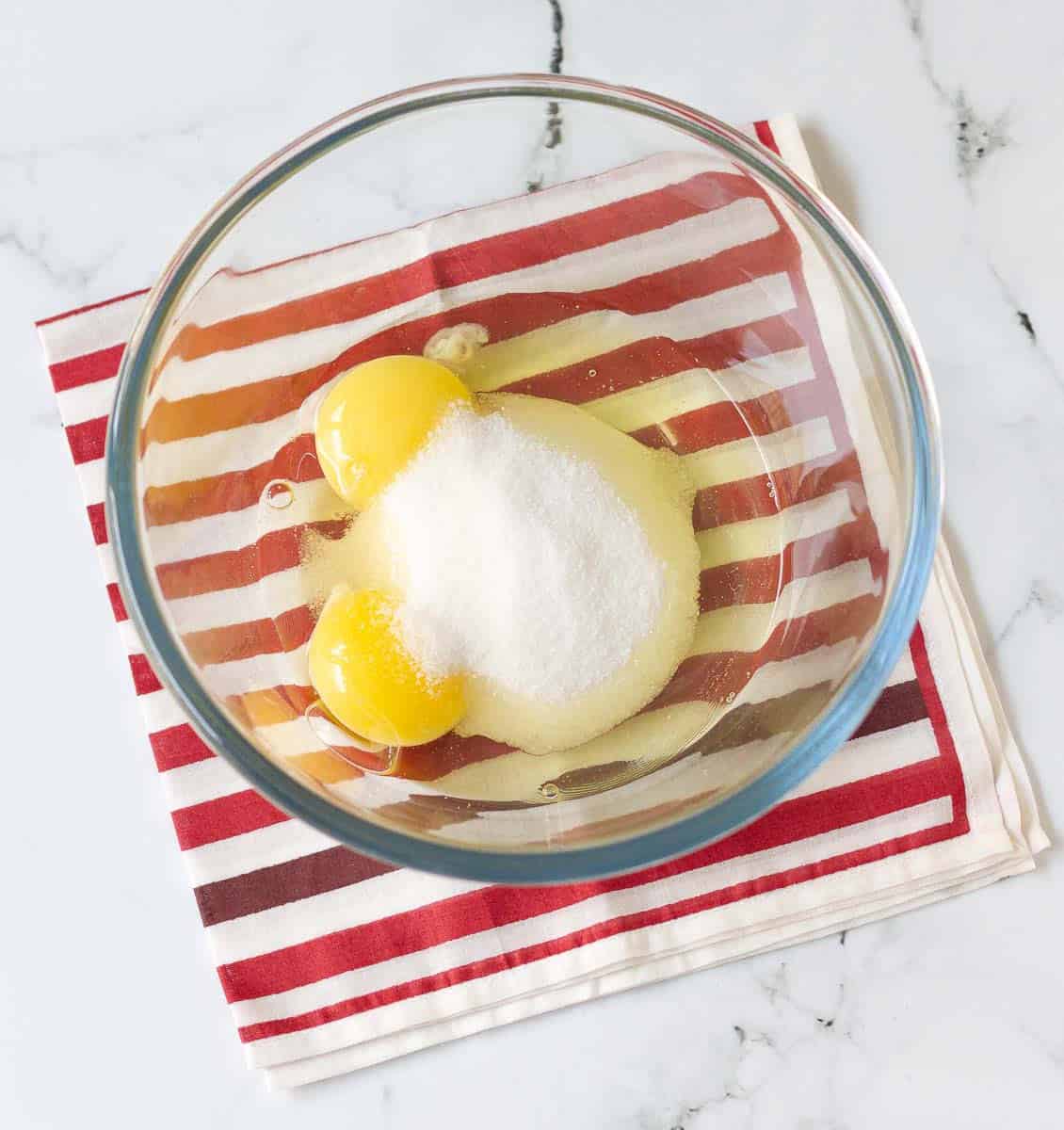 Eggs and sugar in a glass mixing bowl on a red striped napkin.