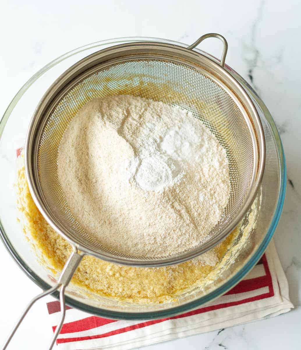 A sieve with flour, baking powder and baking soda on top of a bowl with egg mixture.