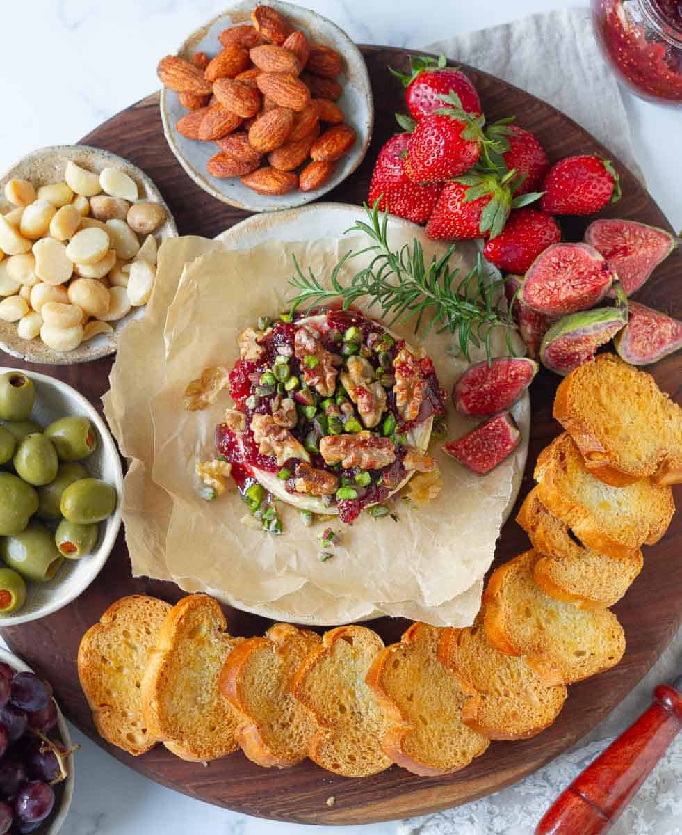 Baked Brie with Fig Jam on a wooden board with almonds, olives, macadamia nuts, strawberries, fresh figs, crostini