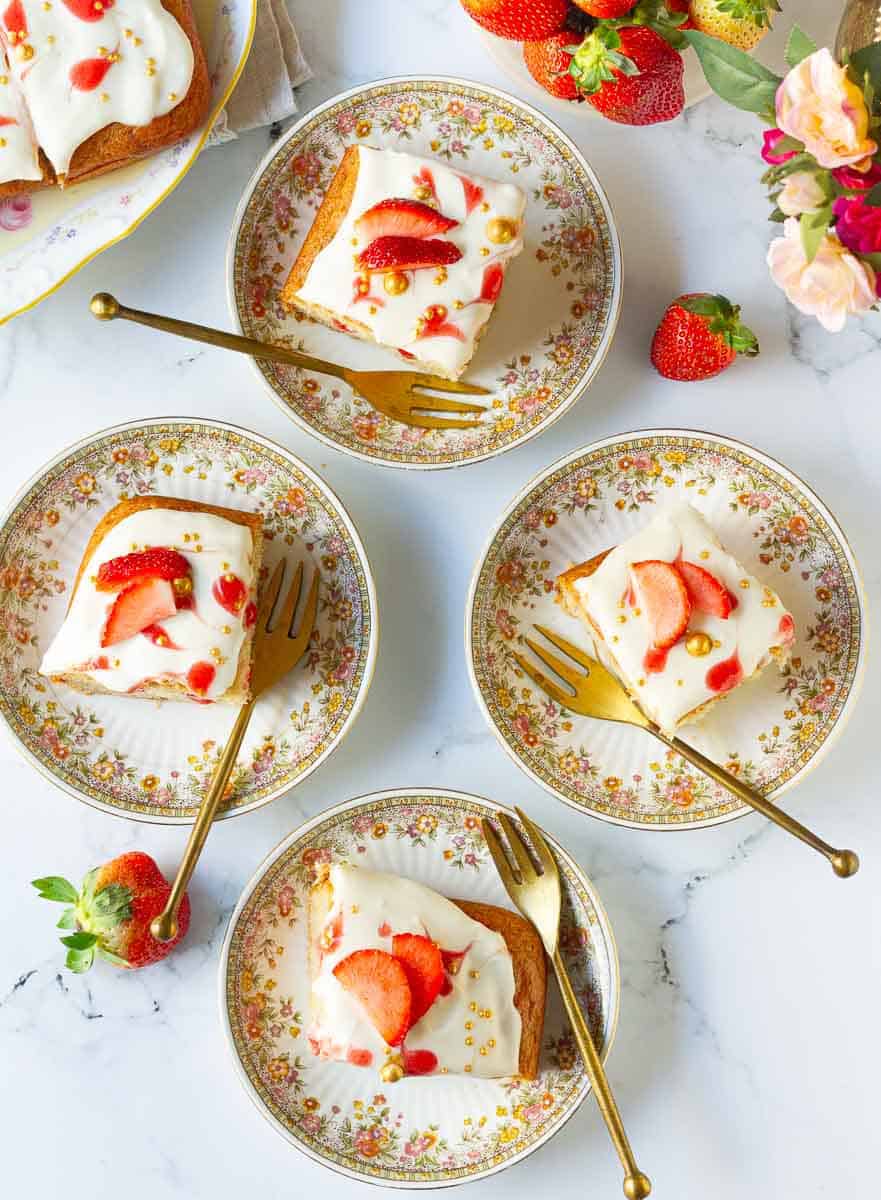 4 squares of poke cake on 4 small dessert plates with forks and fresh strawberries.