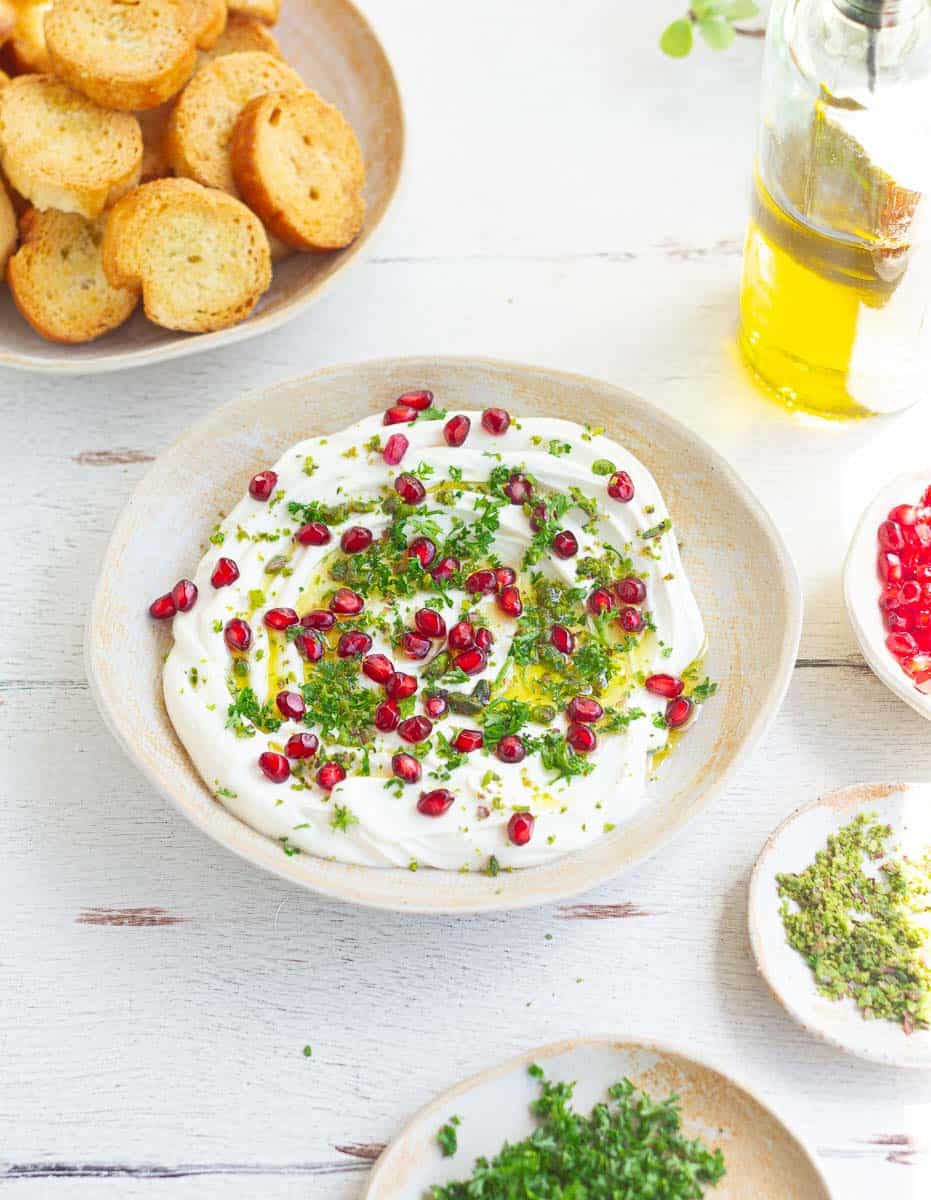 Creamy dip topped with parsley, pomegranates with crostini and other ingredients around.
