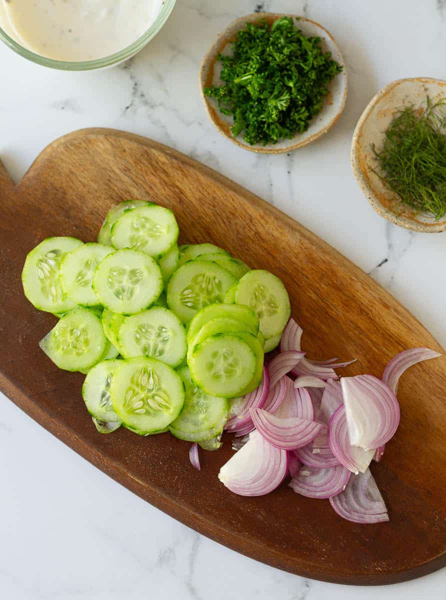 sliced cucumber and onions on a wooden chopping board. Parsley and dill in small bowls