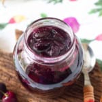A jar with cherry jam on a wooden surface with a spoon on the side.