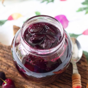 A jar with cherry jam on a wooden surface with a spoon on the side.
