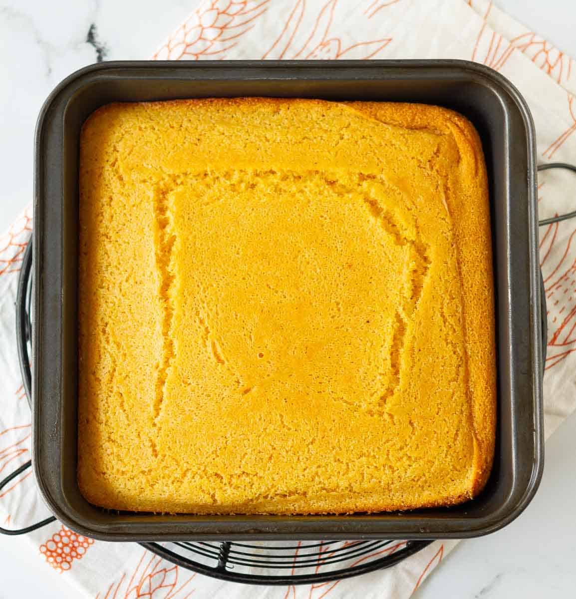 Baked cornbread in a square metal baking pan on a trivet and kitchen towel.
