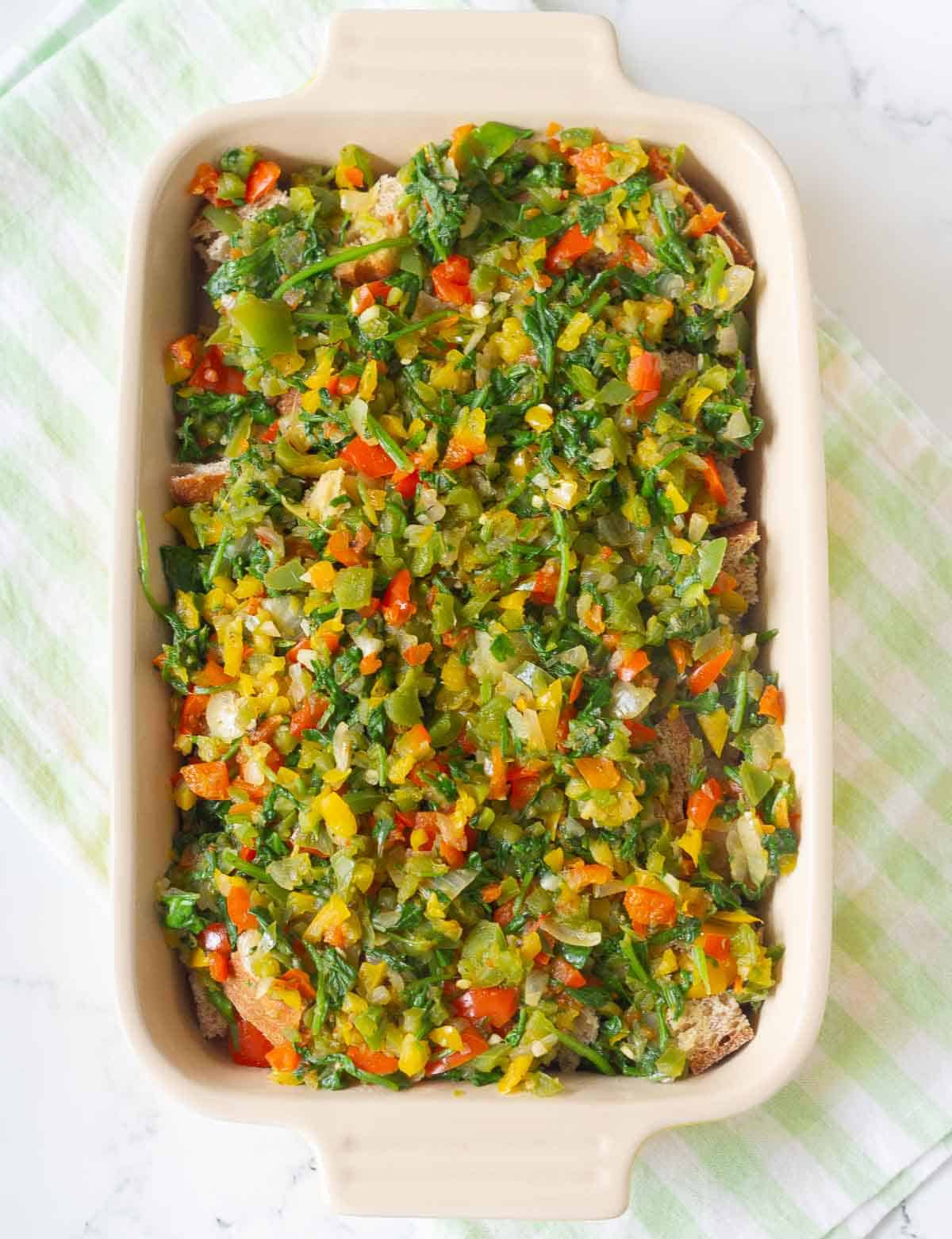 Cooked veggies layered on top of the cubed bread in a casserole dish.