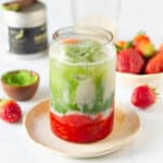 strawberry matcha latte in a glass on a small plate with ingredients in the background.