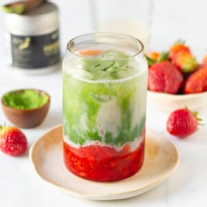 strawberry matcha latte in a glass on a small plate with ingredients in the background.