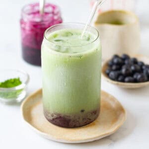 Blueberry matcha latte in a glass with blueberry syrup and blueberries in the background.