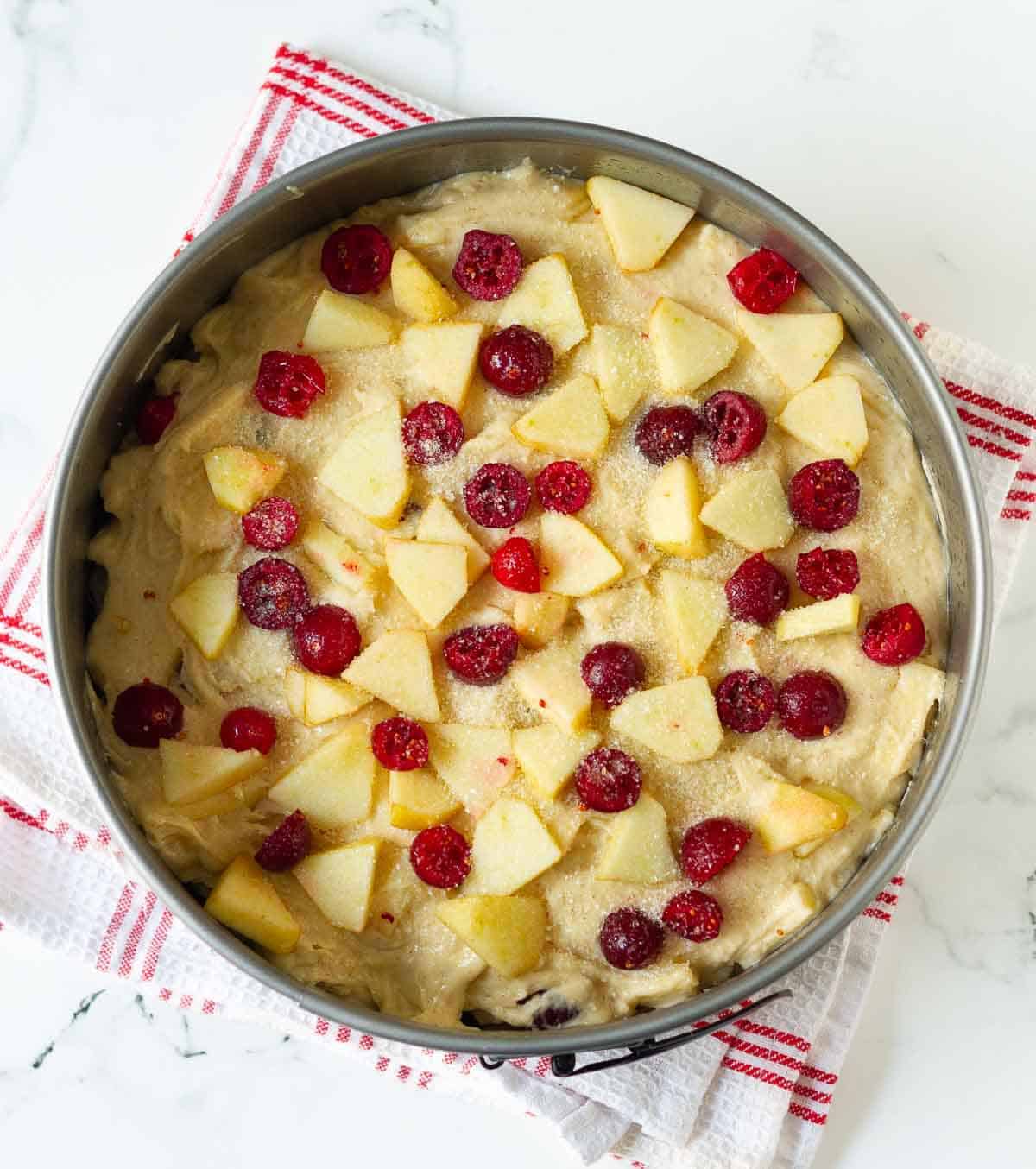 Cake batter in a cake pan with apples and cranberries on top.