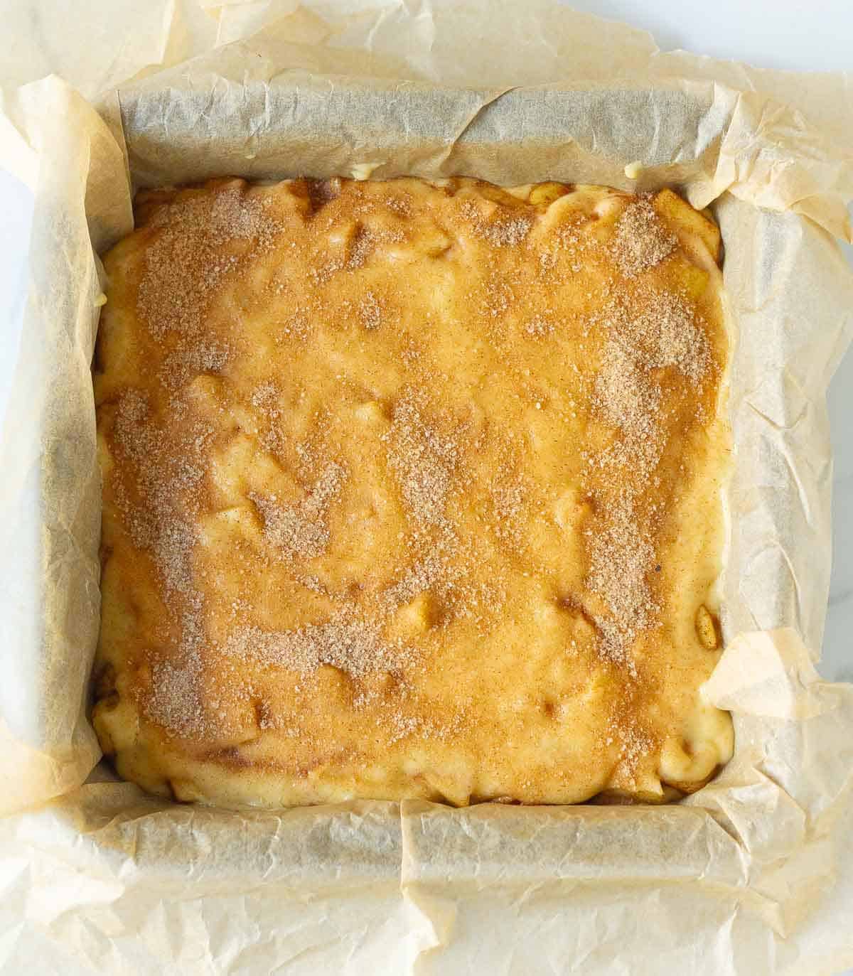 cake batter in a parchment lined cake pan with cinnamon sugar on top.