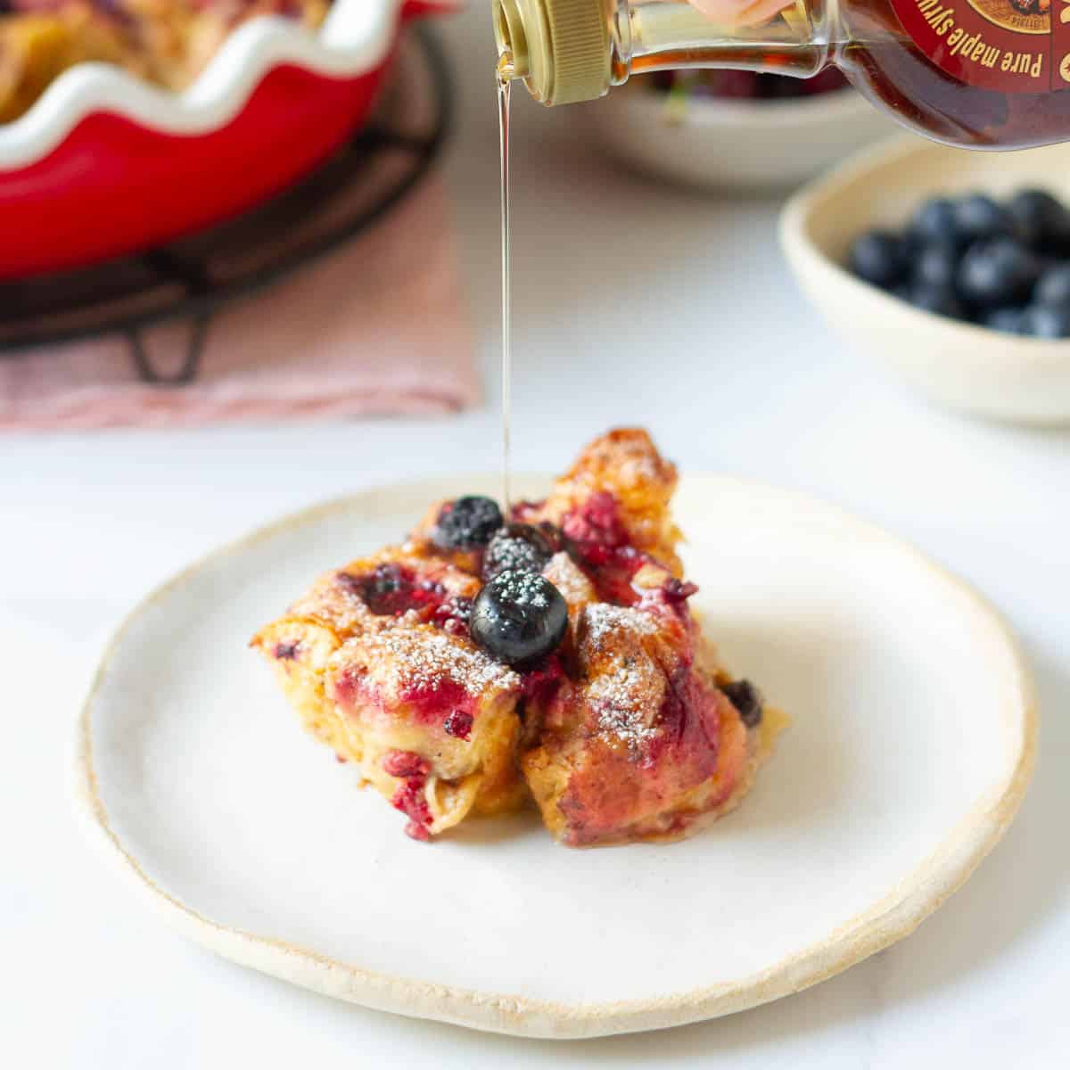 A piece of croissant casserole on a small plate getting a drizzle of maple syrup.