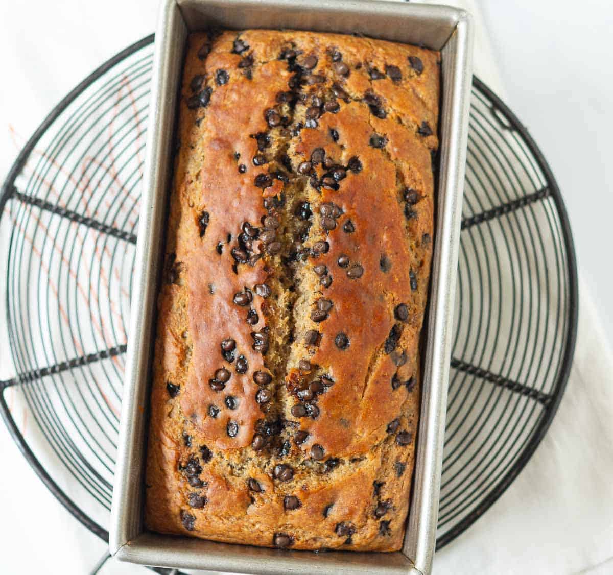 Baked chocolate chip banana bread in a loaf pan on a metal trivet.