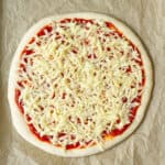 Rolled out pizza dough topped with tomato sauce and grated cheese on parchment paper.