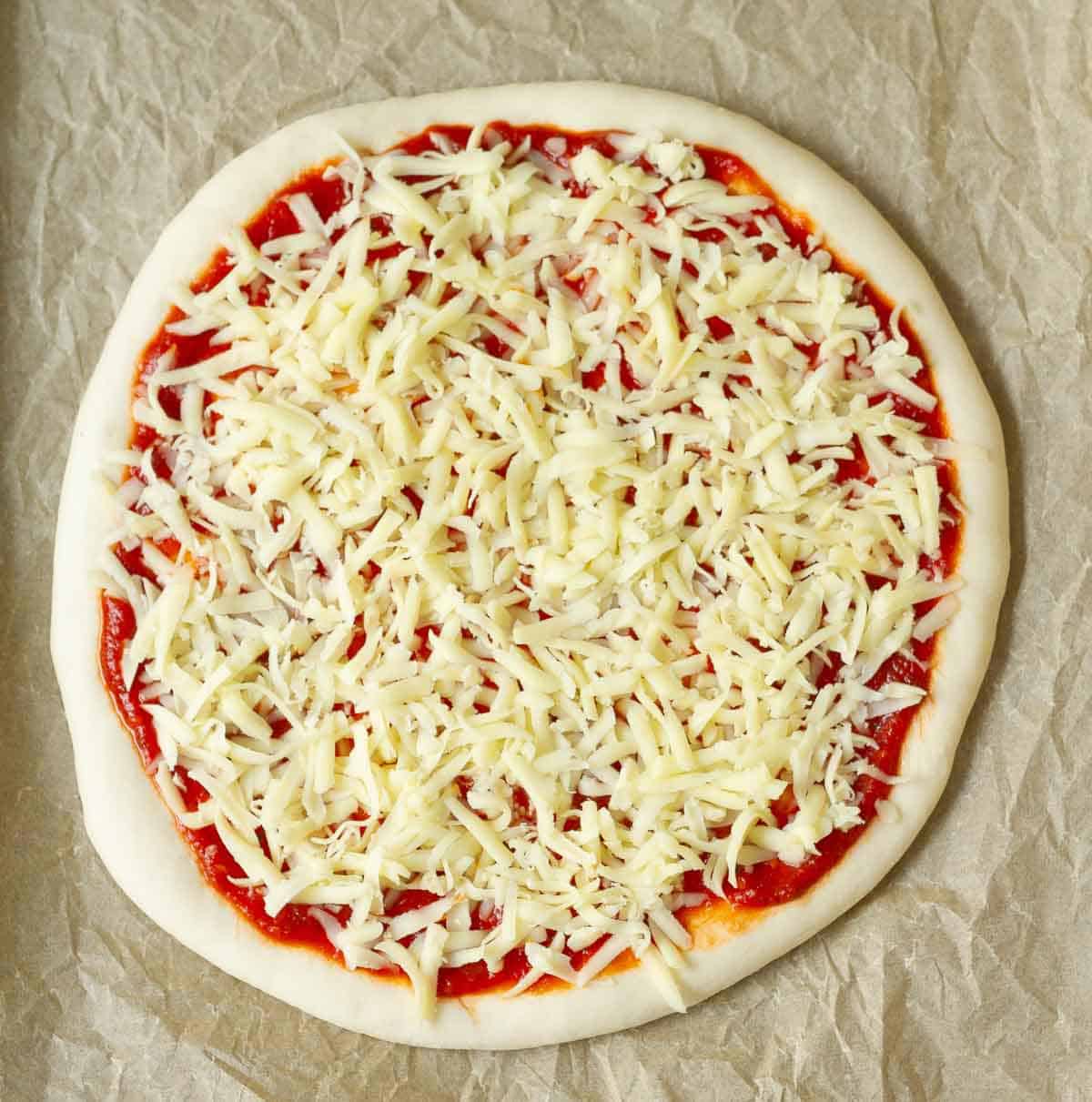 Rolled out pizza dough topped with tomato sauce and grated cheese on parchment paper.