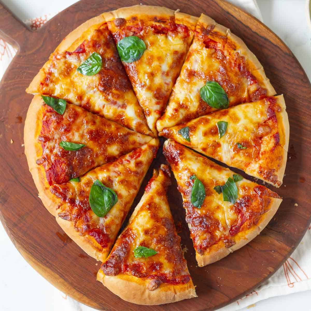 Slice cheese pizza on a wooden board with basil leaves on top.